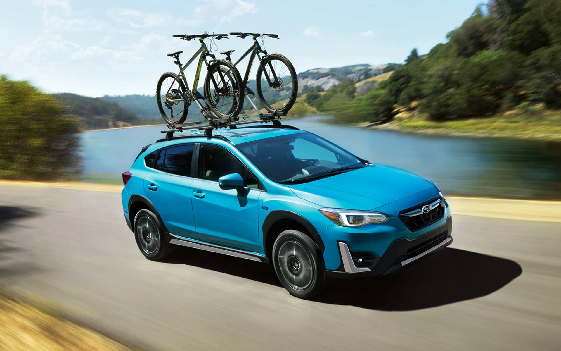 A blue Crosstrek Hybrid with two bicycles on its roof rack driving beside a river | Subaru Superstore of Chandler in Chandler AZ